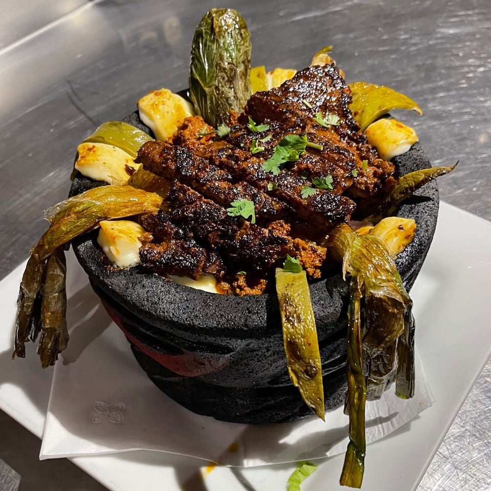 A black stone bowl filled with grilled meat, cheese cubes, and green peppers on a white plate.
