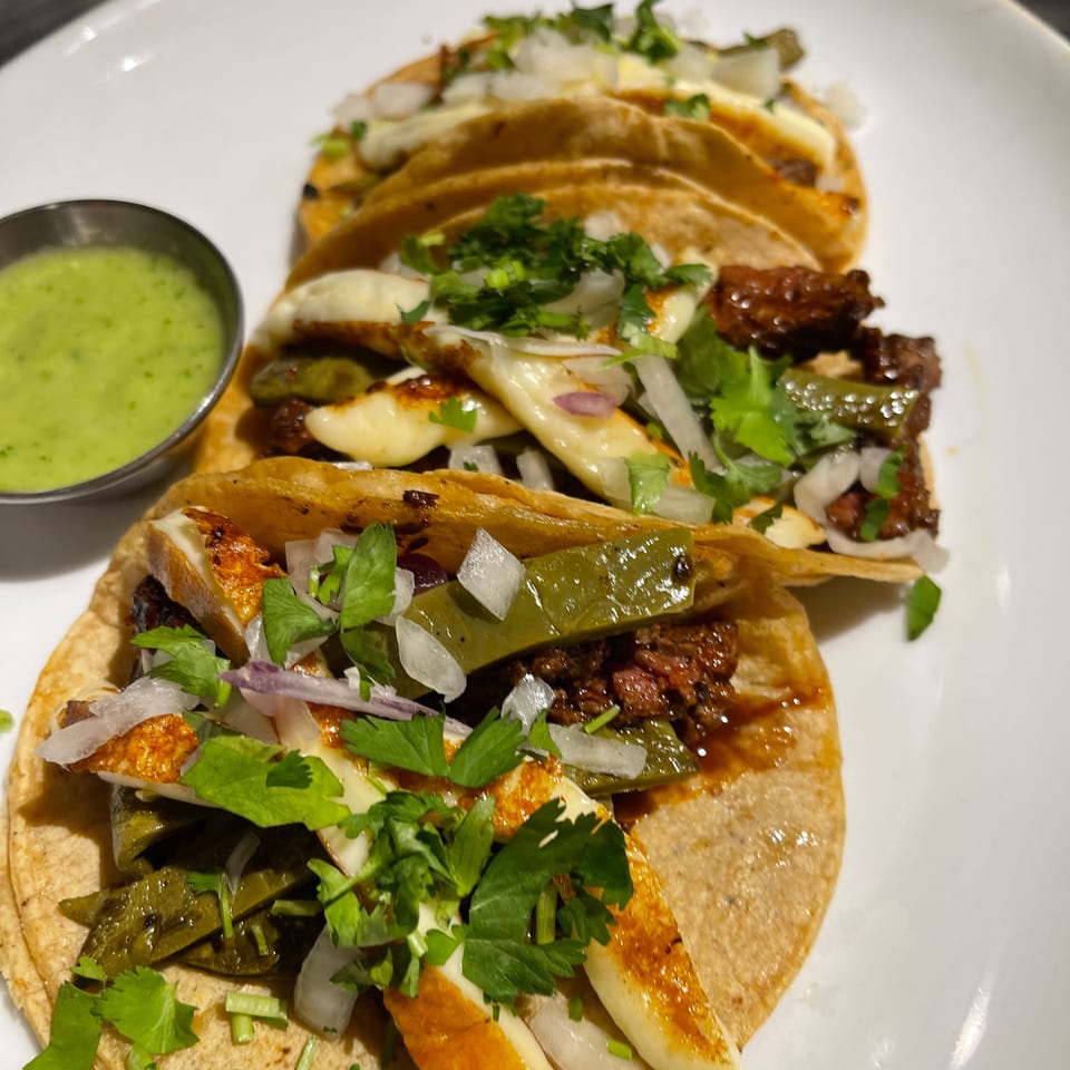 Three tacos with grilled cheese, meat, peppers, onions, and cilantro, served with green sauce.